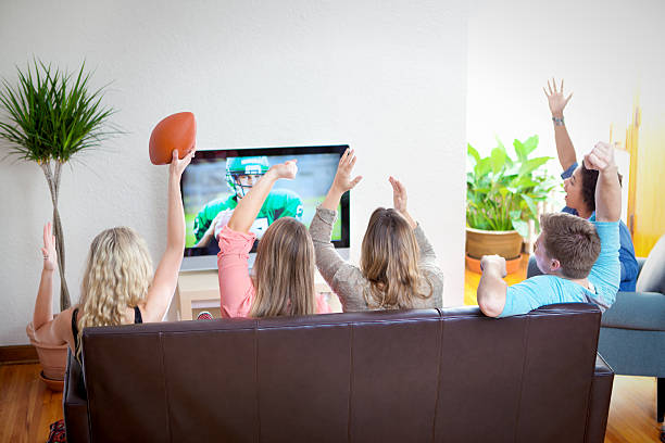 A group of young friends watching a sports football game together at home. The excited teenager or college student spectator audience sits on a living room sofa and chair, cheering and holding a ball. Girls and a boy enjoy a broadcast program. The image on TV screen is the photographer’s.